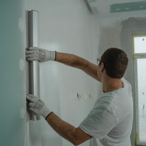 A man in a white shirt and gloves using a metal ruler for indoor wall renovation work.
